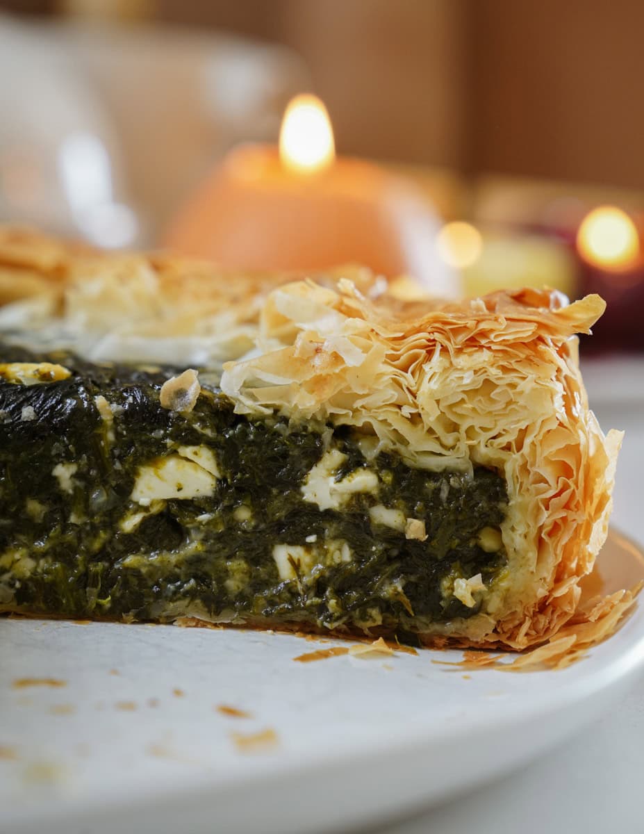 Close-up of a sliced spinach and feta pie with crispy, golden phyllo pastry. The pie is on a white plate, and blurred candle lights glow softly in the background, adding a cozy atmosphere.