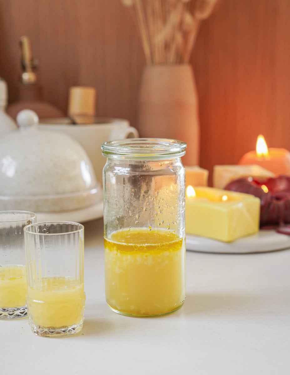 A jar and two glasses contain a yellow liquid, possibly clarified butter, on a kitchen counter. Behind them are butter blocks, candles, and decorative items, set against a warm-toned background.