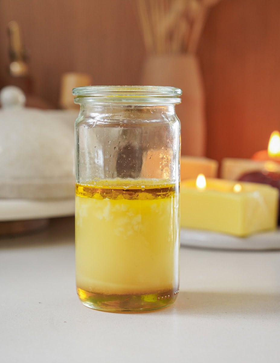 A clear jar containing separated layers of melted butter sits on a white countertop. The background features a butter dish, burning candles, and a room with warm lighting, creating a cozy ambiance.