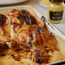 A roasted spatchcock chicken with golden, crispy skin on a baking tray. The chicken is seasoned and surrounded by juices. In the background, theres a jar of mustard, slightly blurred.