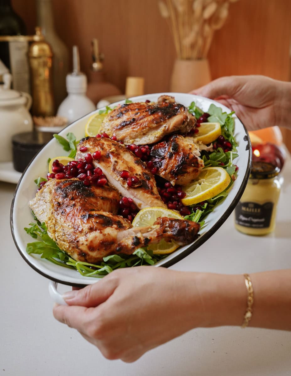 A person holds a platter of grilled chicken garnished with lemon slices, pomegranate seeds, and laid on a bed of greens. Various kitchen items and a jar of mustard are visible in the background.