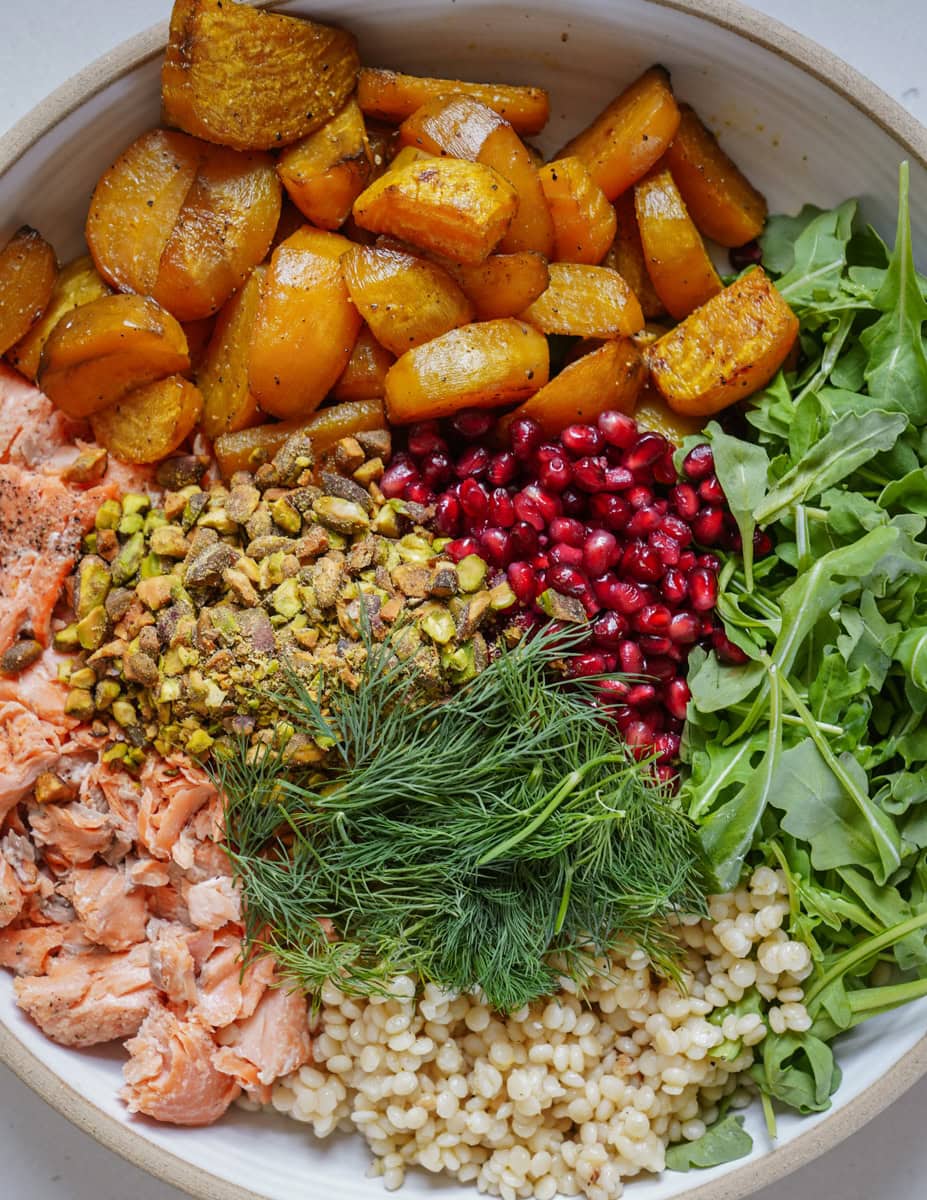 A bowl filled with roasted orange vegetables, arugula, pomegranate seeds, dill, crushed pistachios, cooked barley, and flaked salmon, artfully arranged in sections.