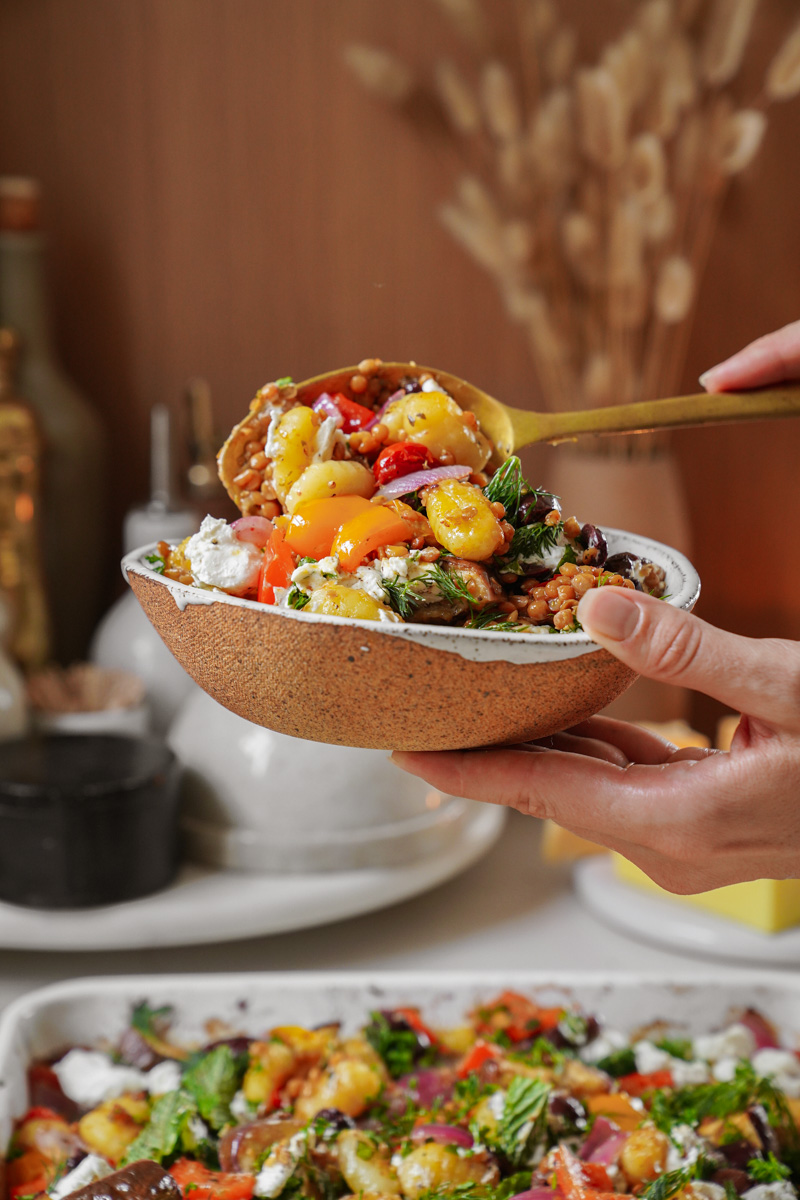 A person holds a bowl filled with a vibrant salad, featuring cherry tomatoes, fresh greens, and grains. A spoonful of the salad is being scooped from a dish below. Various kitchen items are blurred in the background.