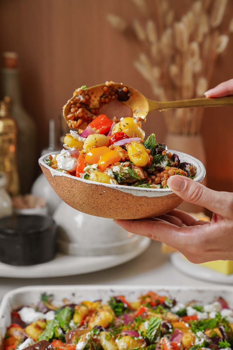 A person holds a ceramic bowl filled with a colorful salad of cherry tomatoes, lentils, and herbs. Theyre using a spoon to serve more salad into the bowl. A tray of ingredients and kitchen items are blurred in the background.