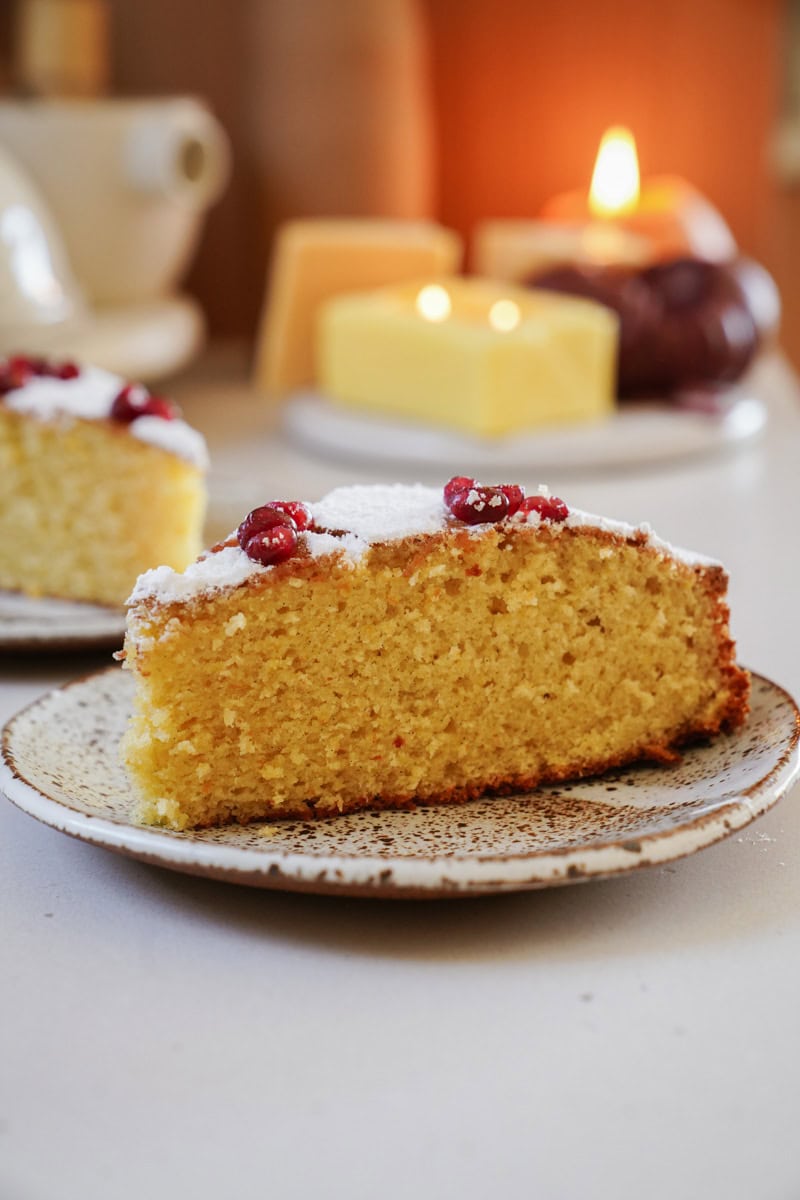 A slice of yellow cake topped with powdered sugar and red berries on a ceramic plate. In the background, theres another slice, a block of butter, and a lit candle on a blurred table.