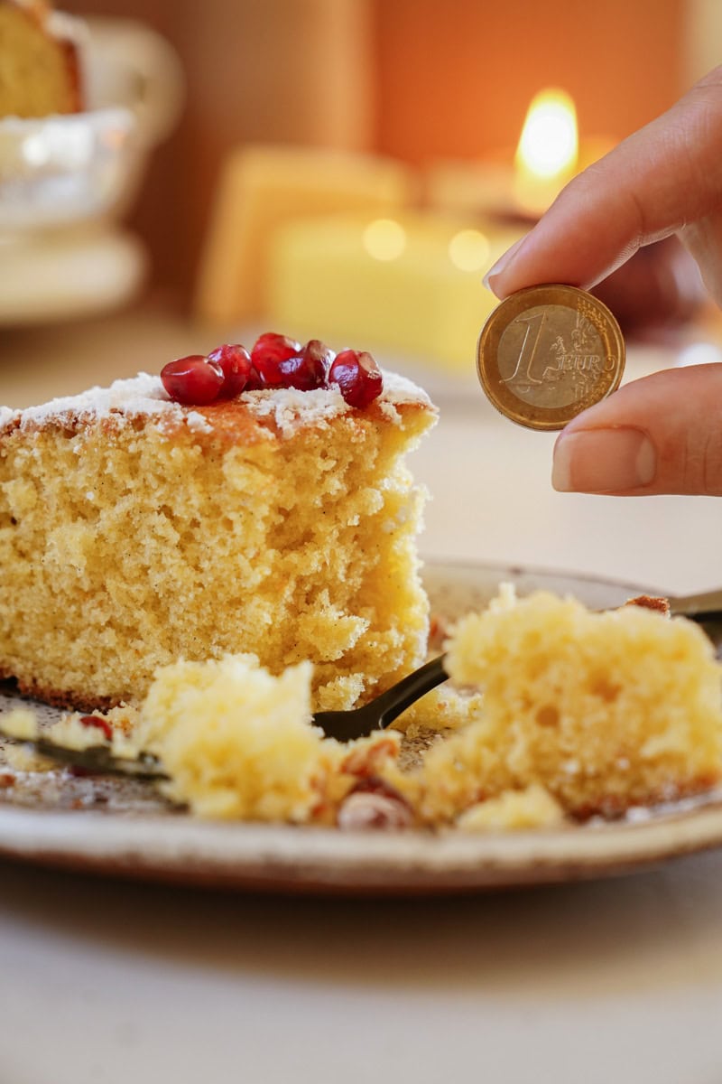 A slice of cake topped with pomegranate seeds on a plate. A hand holds a coin above the cake. Candles and warm lighting in the background.