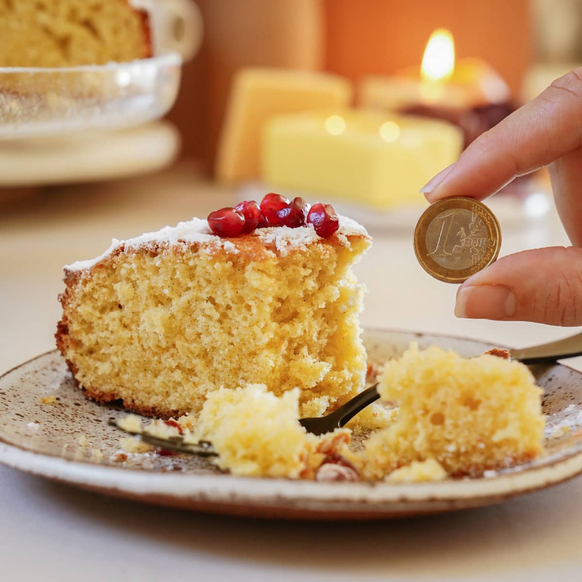 A slice of cake with pomegranate seeds on top sits on a speckled plate. A hand is holding a one euro coin near the cake. A fork is on the plate with crumbs scattered around. Candles and a blurred background create a cozy atmosphere.